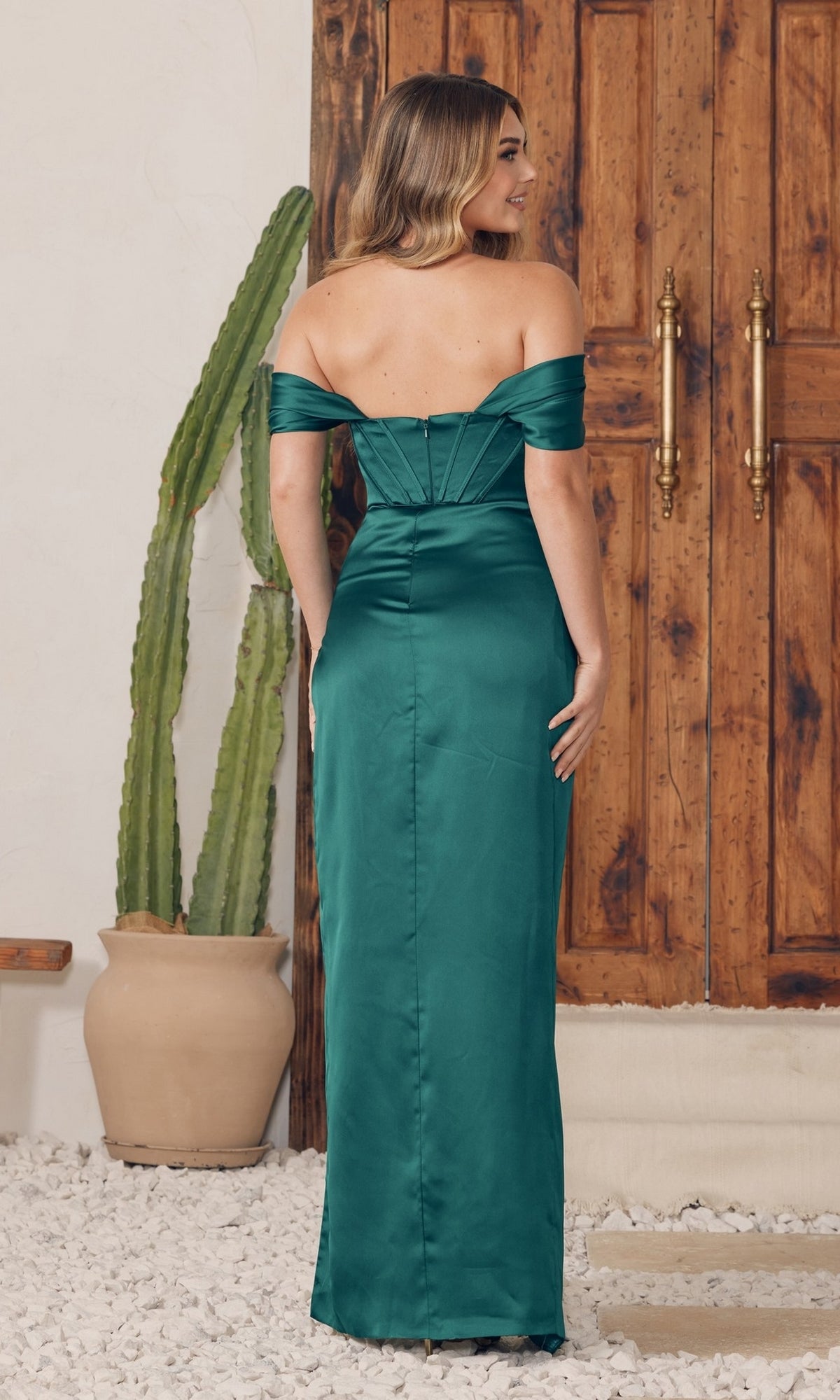 A woman in the Off-the-Shoulder Sleek Long Prom Dress R1236 stands facing a wooden door, with a tall potted cactus beside her on a stone floor.